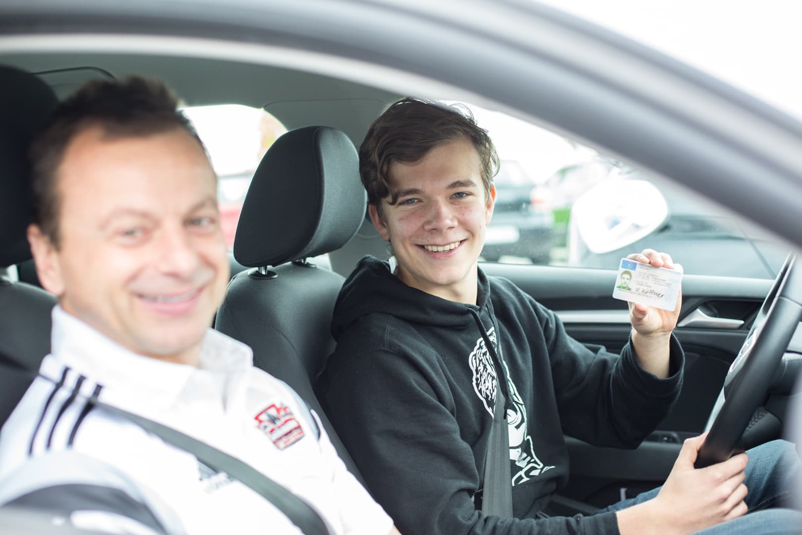 Fahrschüler zeigt lachend seinen neuen Führerschein, daneben sein Fahrlehrer